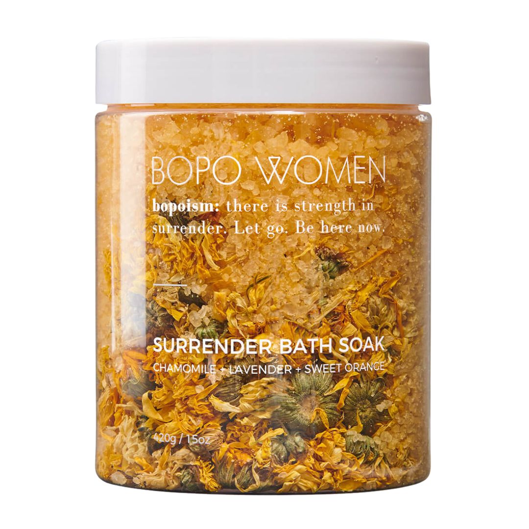 A clear plastic jar of Bopo Women 'Surrender' bath soak with a white lid, filled with dried chamomile, lavender, and sweet orange botanicals, on a white background.